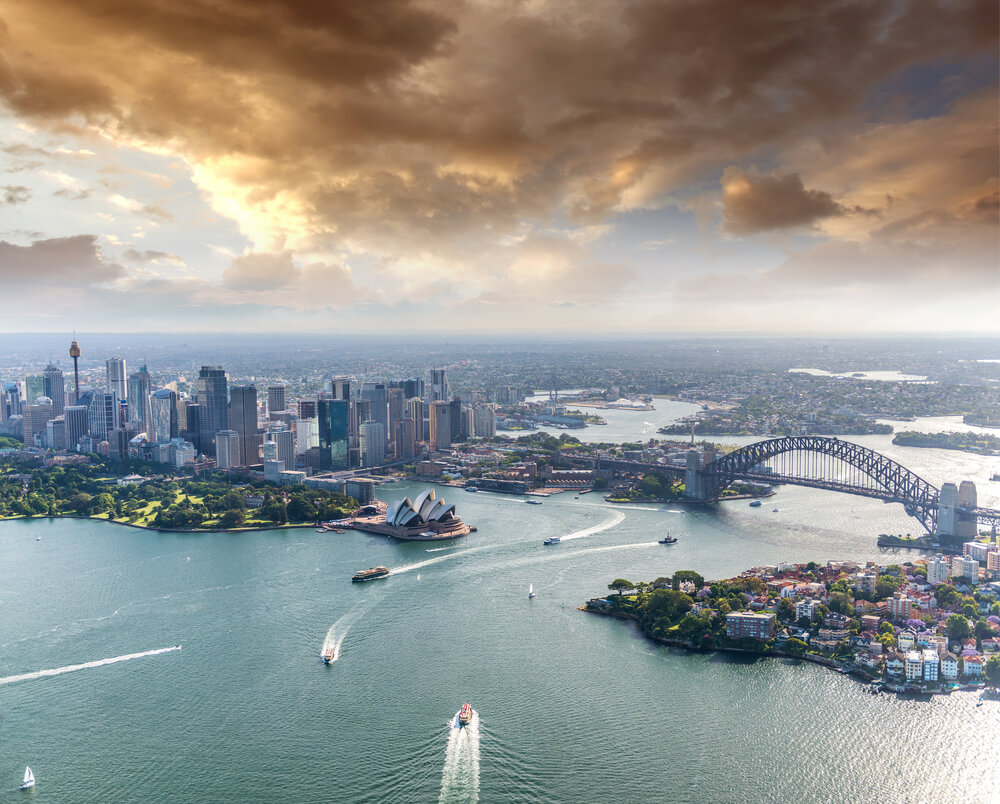 Wide view of bustling Sydney CBD, Story Bridge and Paramatta River. Cloudy sky and high rises on the left and suburbs on the right. Gladesville Plumbing Services also services Sydney CBD.
