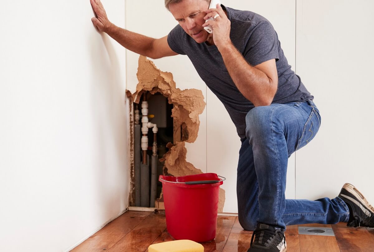 GPS Services - Leaking Pipes Emergency Plumber Man calling professional plumber in North Sydney, kneeling over red bucket catching water from a leaky pipe. Wall next to him is broken from water damage. Services available for leaky toilet, tap and showers too.