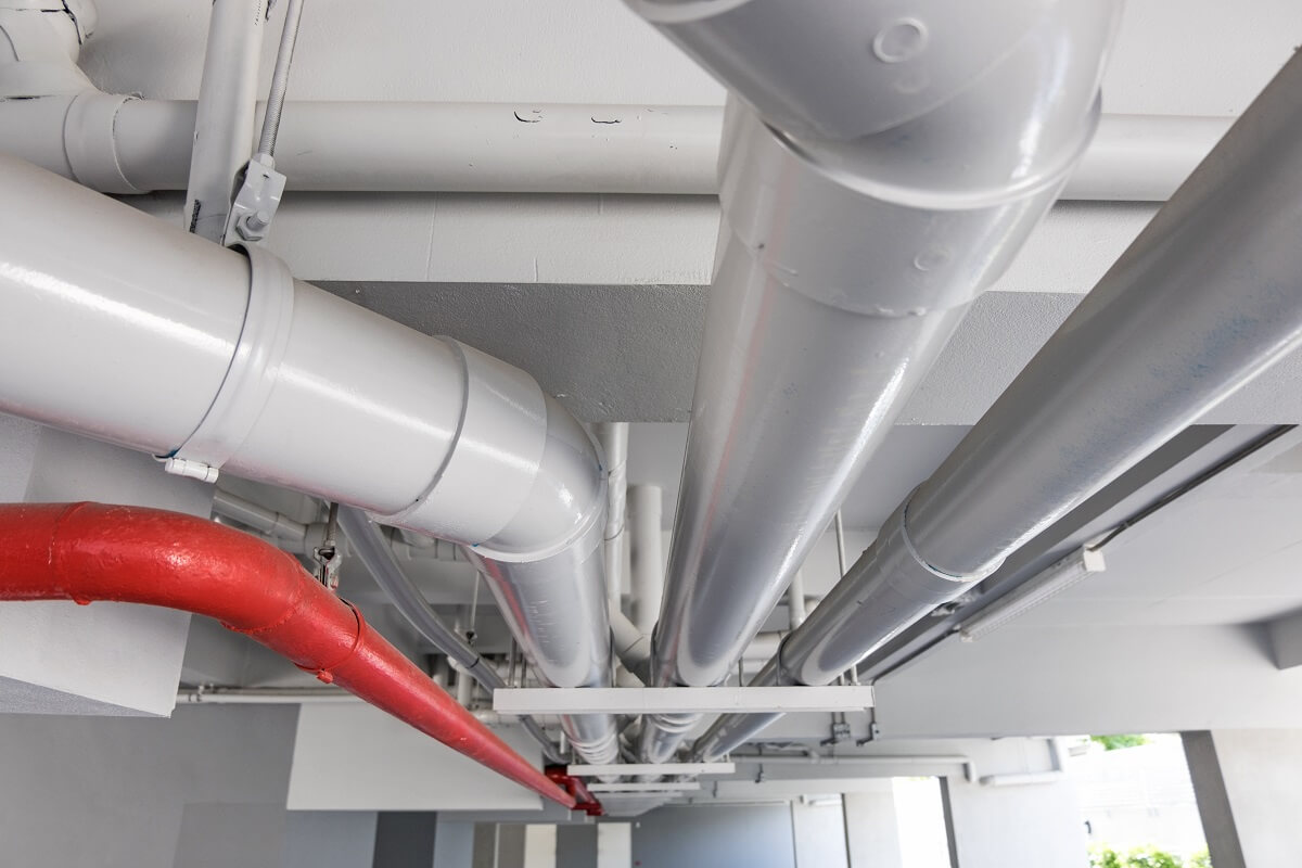 Three large and thick white pipes and one red pipe along ceiling of parking garage. Professional commercial plumbing for body corporate, strata properties, apartment buildings and agency plumbing for North Sydney.