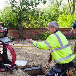 Two professional plumbers in hi vis uniform pulling through red CCTV cord from a reel down into underground system. Plumbing services in North Sydney.