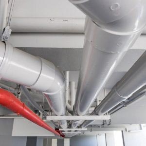 Three large and thick white pipes and one red pipe along ceiling of parking garage. Professional commercial plumbing for body corporate, strata properties, apartment buildings and agency plumbing for North Sydney.